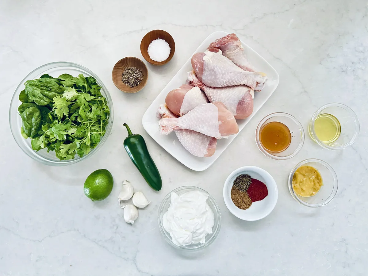 A flat lay of ingredients for Air-Fried Peruvian Chicken with Green Crema on a white surface, featuring raw chicken drumsticks, leafy greens, jalapeño, lime, garlic, spices, salt, pepper, oil, honey, mustard, and a bowl of yogurt.