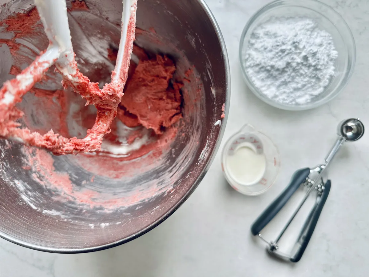 Overhead view of a mixing bowl with red batter for Strawberry Shortcake Cookies and a mixer attachment, next to a bowl of powdered sugar, a small cup of milk, and a metal cookie scoop on a white surface.