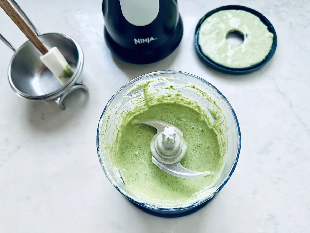 A small blender filled with green sauce for Air-Fried Peruvian Chicken with Green Crema sits on a counter. The lid and a spatula, both smeared with the sauce, rest nearby on the clean, light-colored surface.