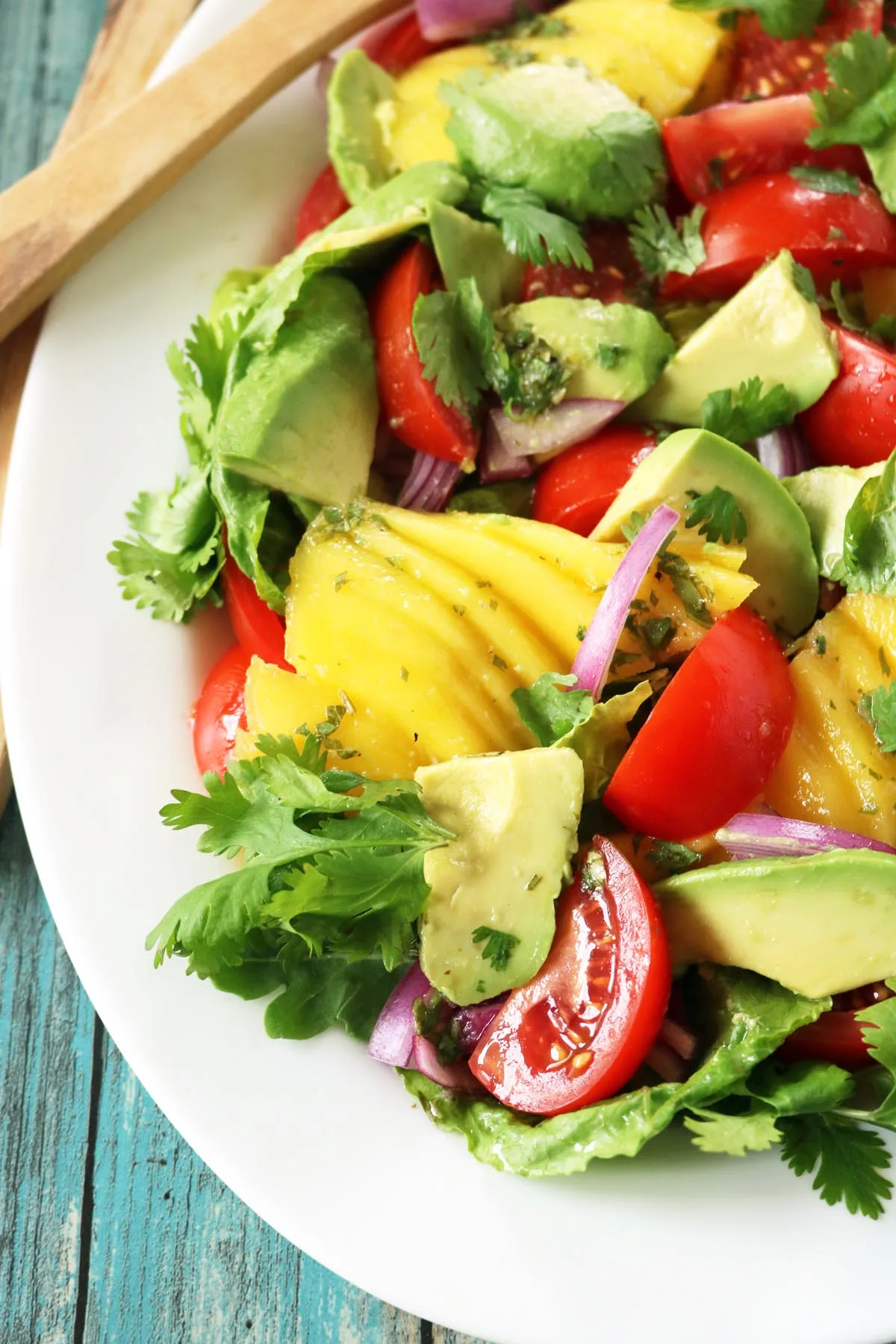 A vibrant Tomato and Avocado Salad featuring ripe mango, tomatoes, avocado, red onion, lettuce, and cilantro on a white plate, beautifully arranged atop a rustic blue wooden table.