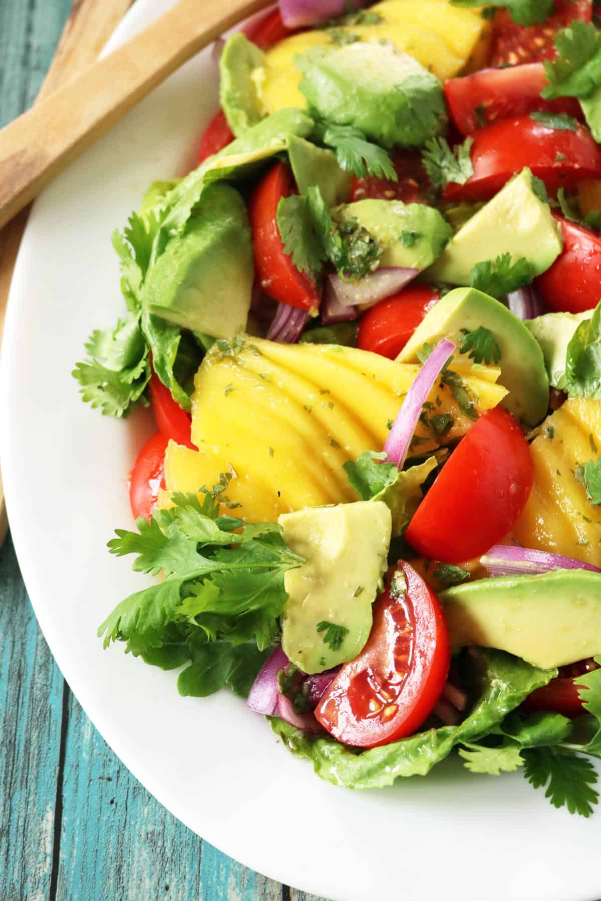 A vibrant Tomato and Avocado Salad featuring ripe mango, tomatoes, avocado, red onion, lettuce, and cilantro on a white plate, beautifully arranged atop a rustic blue wooden table.