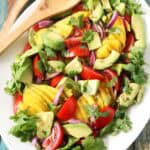 A white platter features a vibrant Mango, Tomato, and Avocado Salad with red onion and cilantro, set on a rustic wooden table beside wooden serving utensils.