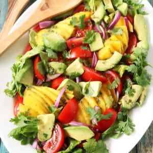 A white platter features a vibrant Mango, Tomato, and Avocado Salad with red onion and cilantro, set on a rustic wooden table beside wooden serving utensils.