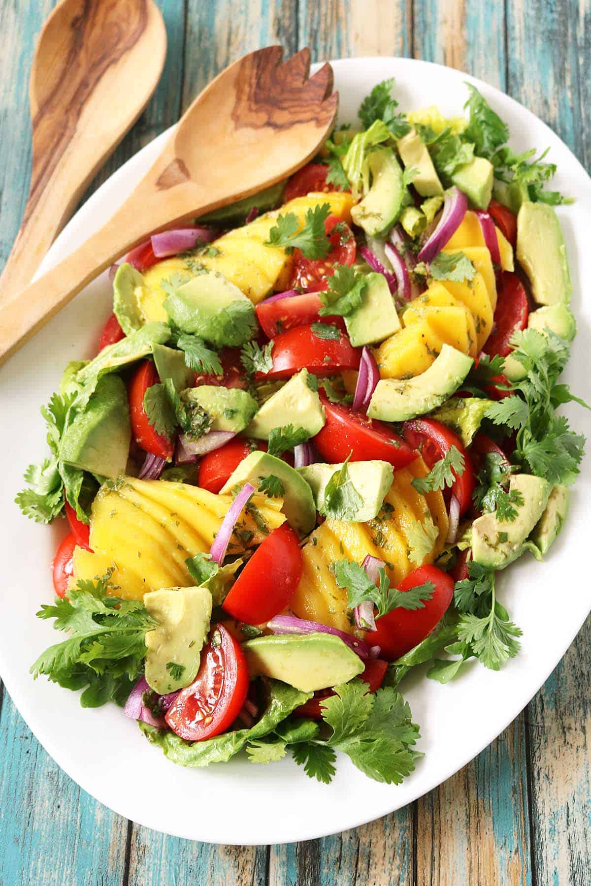 A white platter features a vibrant Mango, Tomato, and Avocado Salad with red onion and cilantro, set on a rustic wooden table beside wooden serving utensils.