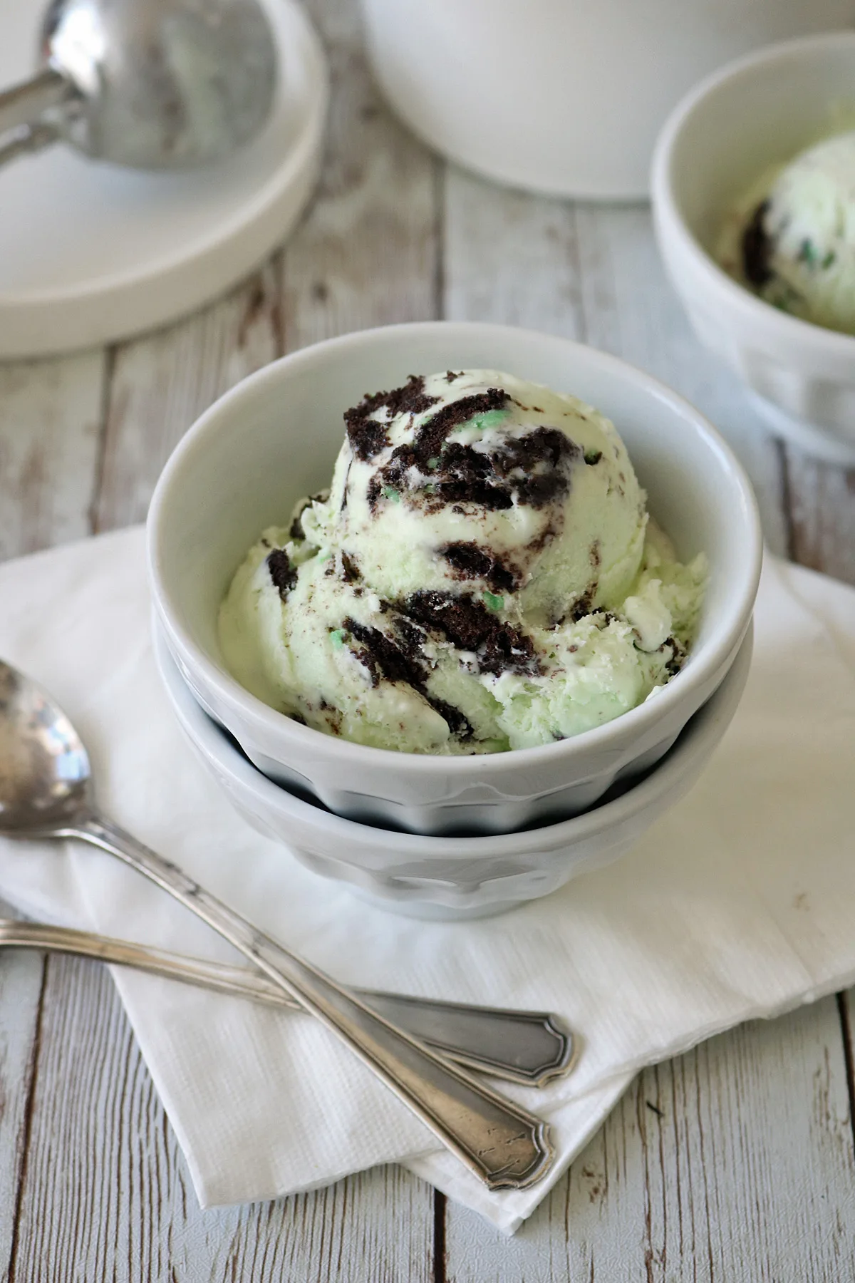 A bowl of mint chocolate chip ice cream with chocolate cookie swirls, placed on a white napkin with two silver spoons beside it on a rustic wooden surface. Another bowl and an ice cream scoop are in the background.