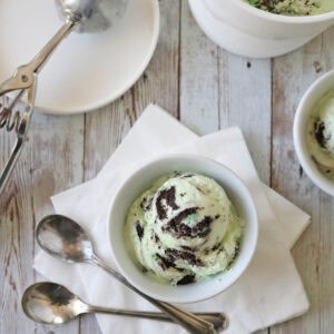 A bowl of mint chocolate chip ice cream sits on white napkins with three spoons beside it. An ice cream scoop and a large container of ice cream are nearby on a rustic wooden surface.