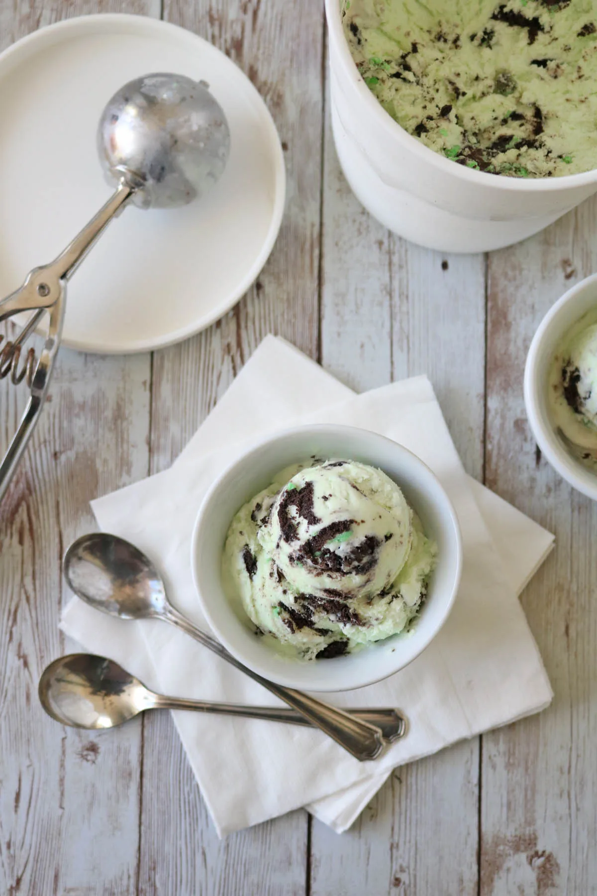 A bowl of mint chocolate chip ice cream sits on white napkins with three spoons beside it. An ice cream scoop and a large container of ice cream are nearby on a rustic wooden surface.