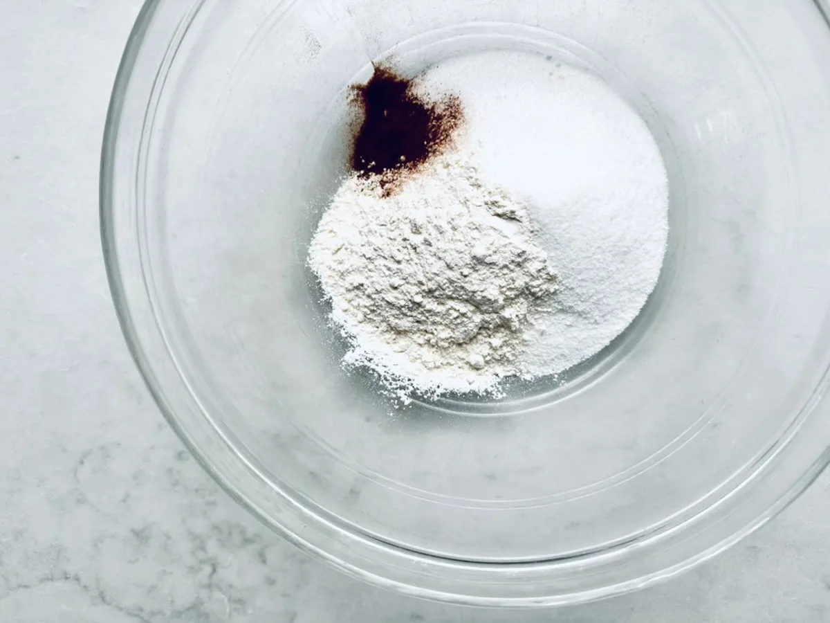 A clear glass bowl containing a pile of flour, a mound of white sugar, and a small amount of brown spice, possibly cinnamon, on a light-colored surface.