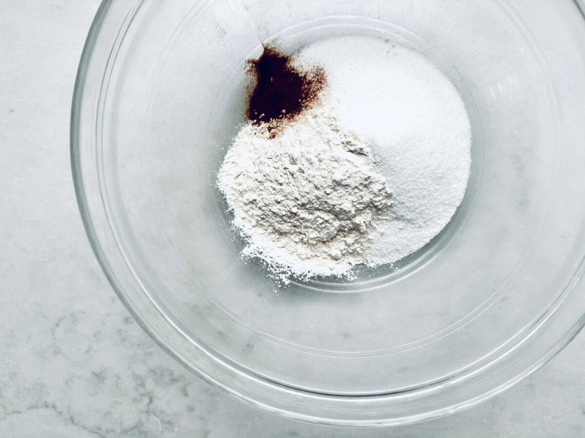 A clear glass bowl containing a pile of flour, a mound of white sugar, and a small amount of brown spice, possibly cinnamon, on a light-colored surface.