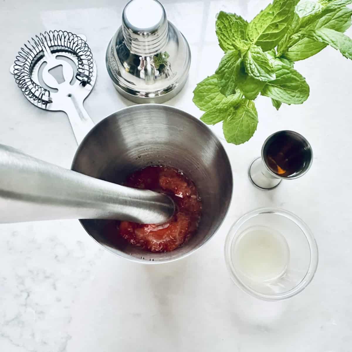 A cocktail shaker with crushed strawberries being muddled, surrounded by a cocktail strainer, jigger with brown liquid, fresh mint, a shaker top, and a glass with clear liquid on a white surface.