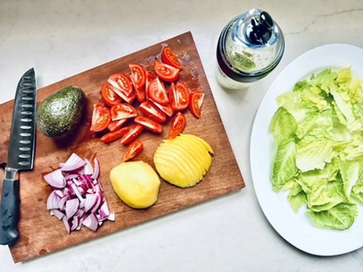 A cutting board with sliced tomatoes, red onion, avocado, and mango for a fresh Tomato and Avocado Salad sits beside a knife; nearby are a plate of lettuce and a jar of salad dressing on a light countertop.