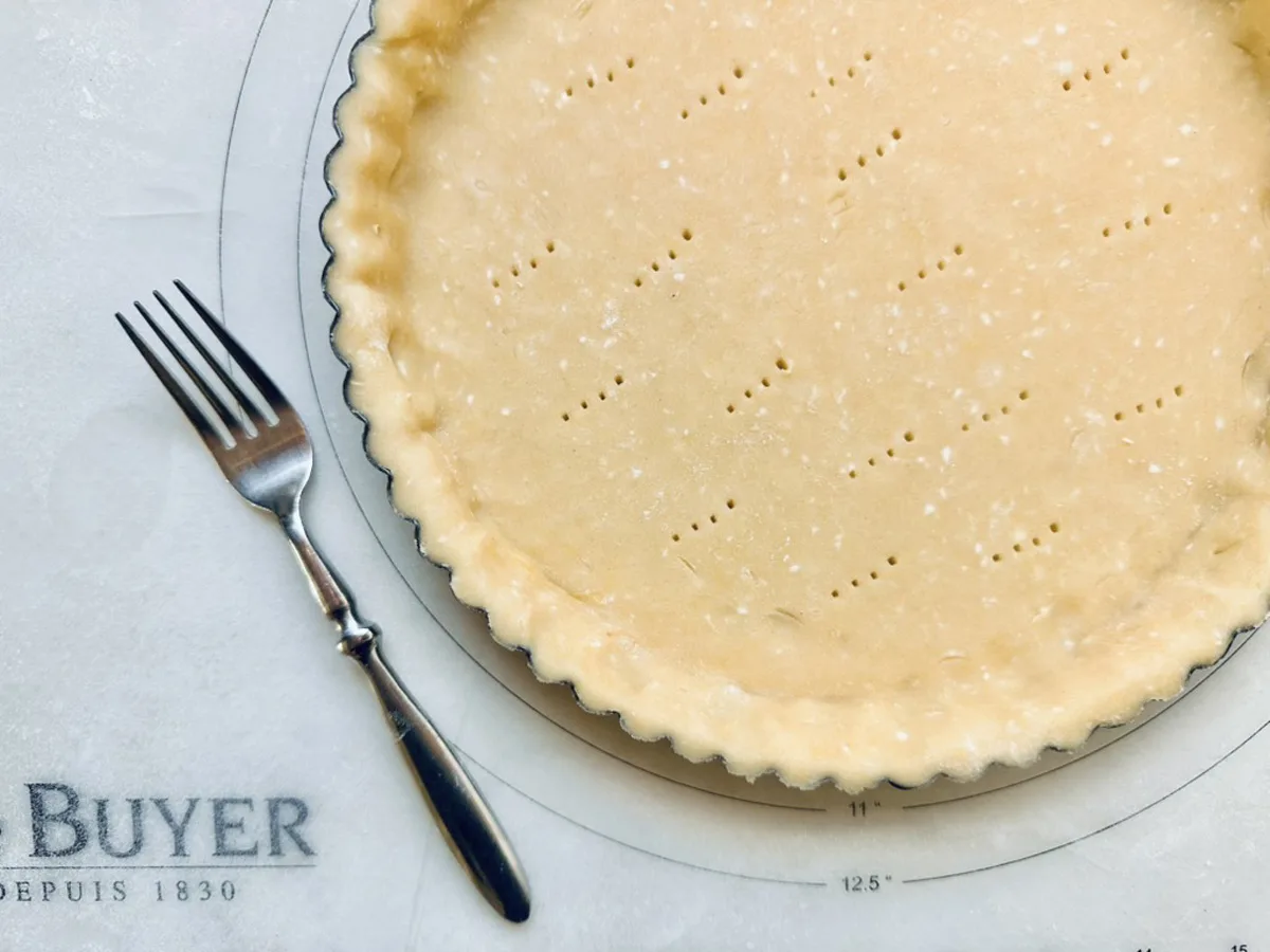 A tart pan filled with raw pie crust, pricked with holes, sits on a pastry mat. A fork rests beside the pan on the left, and part of the mat displays the word BUYER and measurement markings.