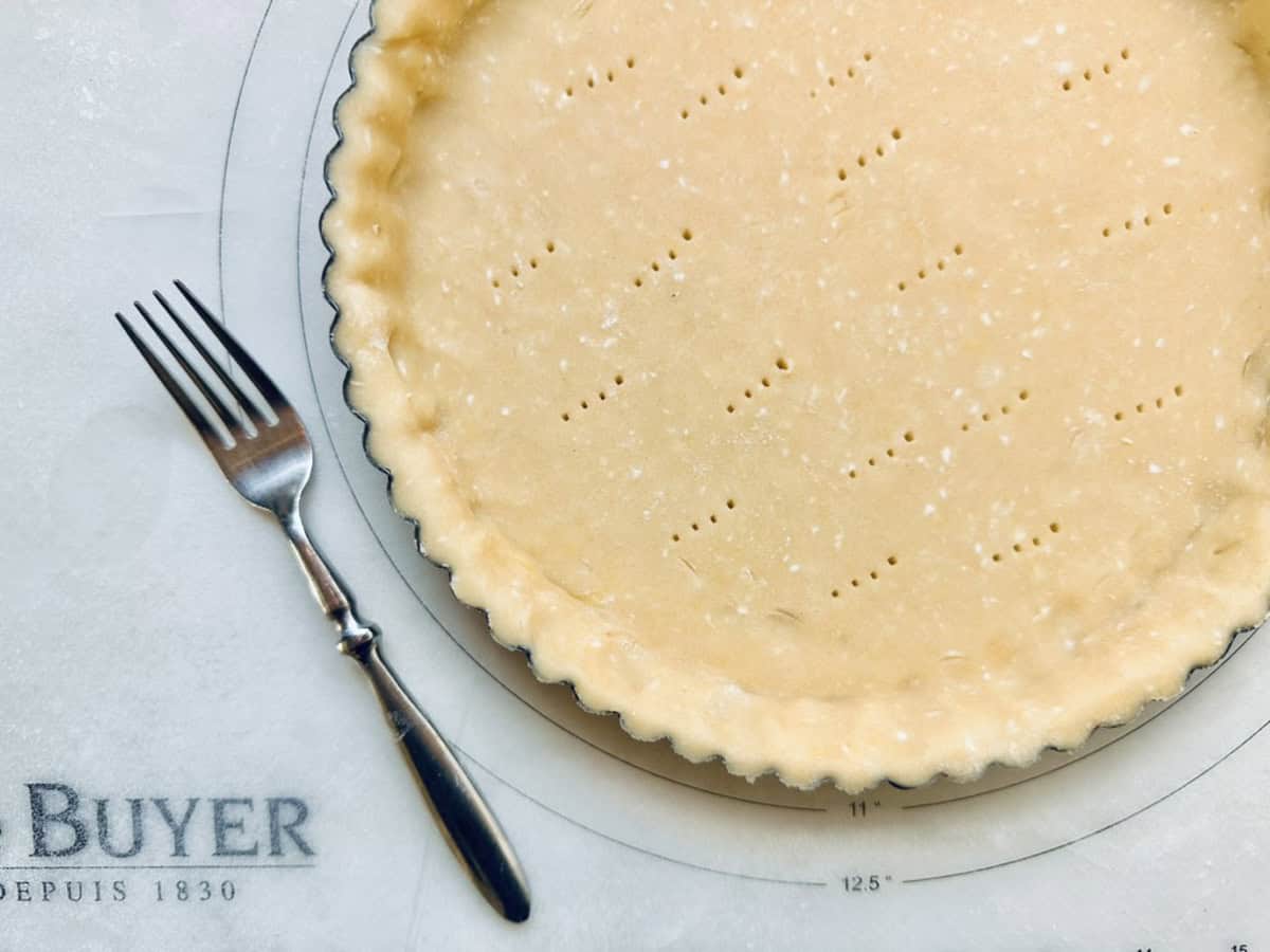 A tart pan filled with raw pie crust, pricked with holes, sits on a pastry mat. A fork rests beside the pan on the left, and part of the mat displays the word BUYER and measurement markings.