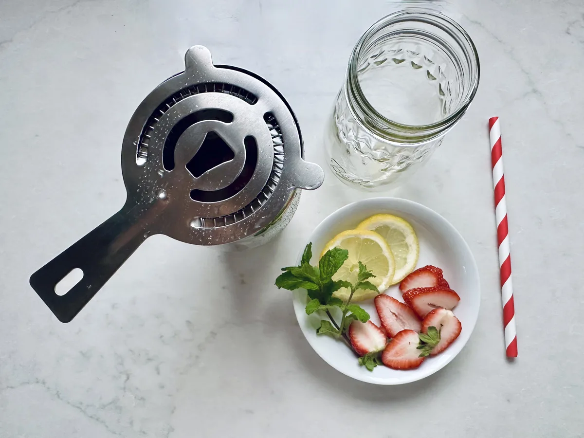 A metal cocktail strainer, an empty mason jar, a red-striped straw, and a small bowl with mint, lemon slices, and sliced strawberries are arranged on a light marble surface.