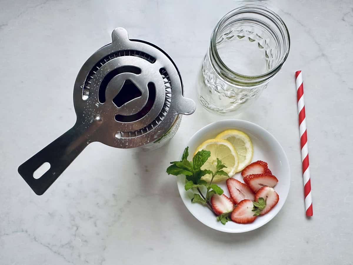 A metal cocktail strainer, an empty mason jar, a red-striped straw, and a small bowl with mint, lemon slices, and sliced strawberries are arranged on a light marble surface.