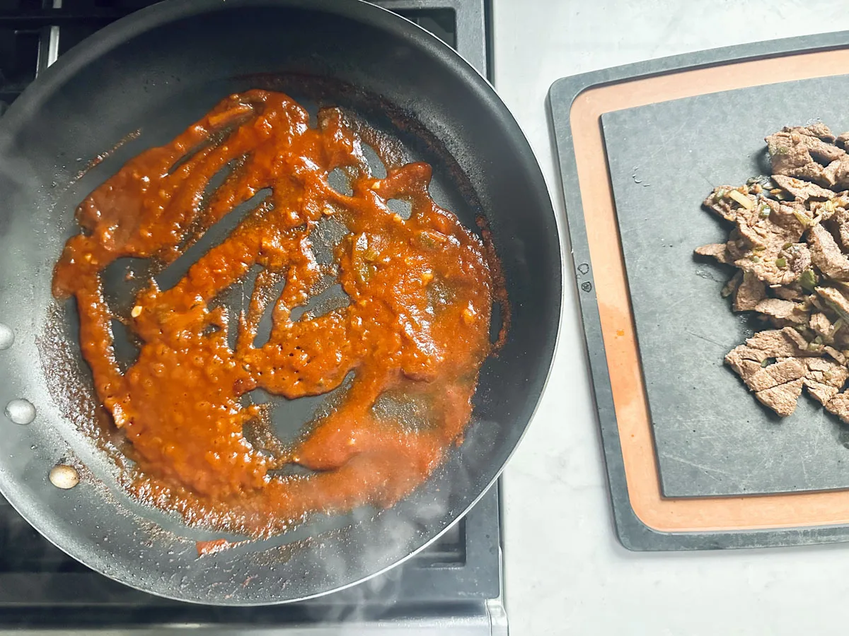 A frying pan with red sauce sits on a stovetop, while cooked sliced beef rests on a black cutting board with a brown border on the countertop nearby.