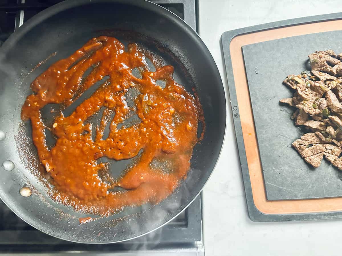 A frying pan with red sauce sits on a stovetop, while cooked sliced beef rests on a black cutting board with a brown border on the countertop nearby.