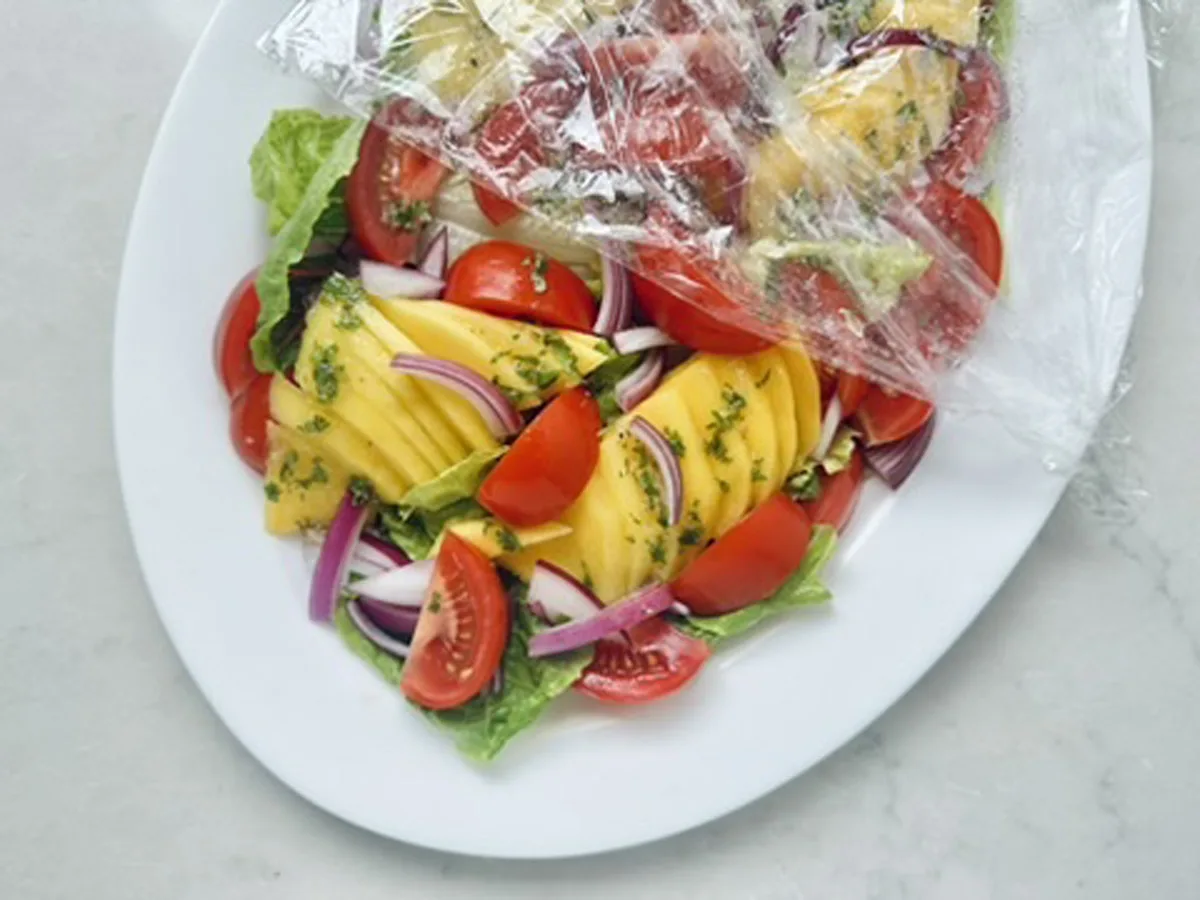 A fresh Mango, Tomato, and Avocado Salad featuring sliced mango, cherry tomatoes, red onion, and lettuce on a white plate, partially covered with clear plastic wrap.