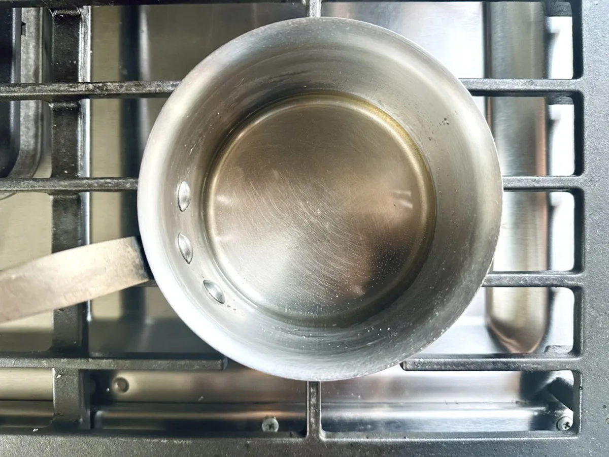 A small, empty stainless steel saucepan sits on a stove with metal grates, ready to prepare ingredients for an Asian Cucumber Ribbon Salad, viewed from above.