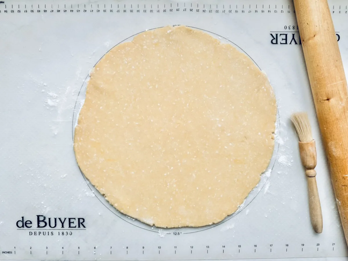A rolled-out round sheet of dough rests on a floured pastry mat, with a wooden rolling pin and a pastry brush placed nearby.
