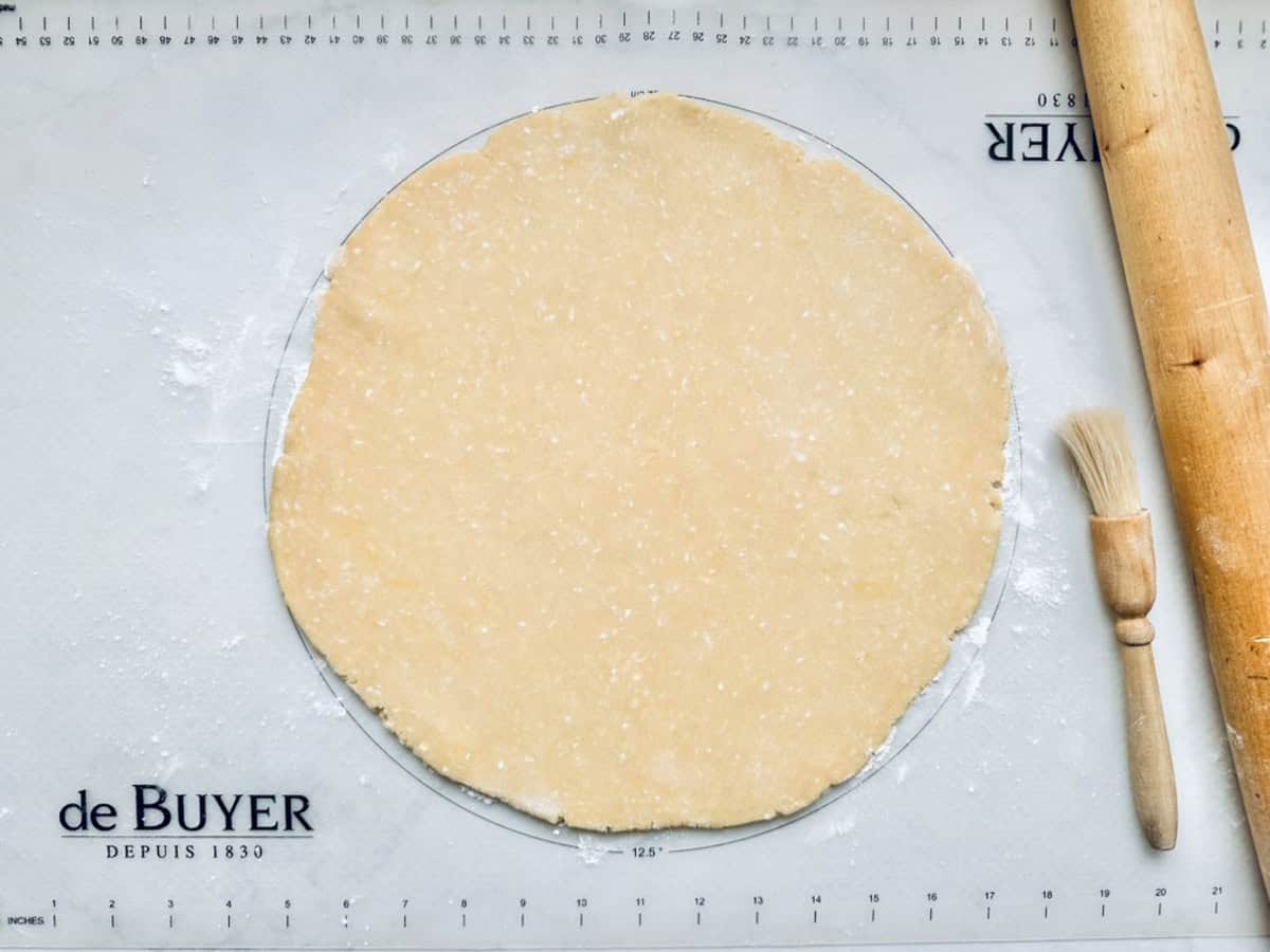 A rolled-out round sheet of dough rests on a floured pastry mat, with a wooden rolling pin and a pastry brush placed nearby.