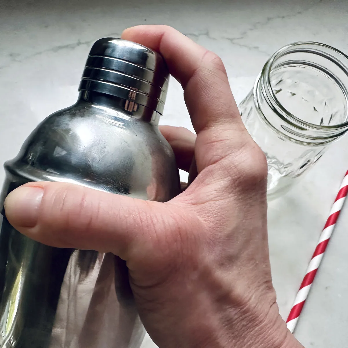 A hand holding a metal cocktail shaker next to an empty glass jar and a red-and-white striped straw on a light surface.