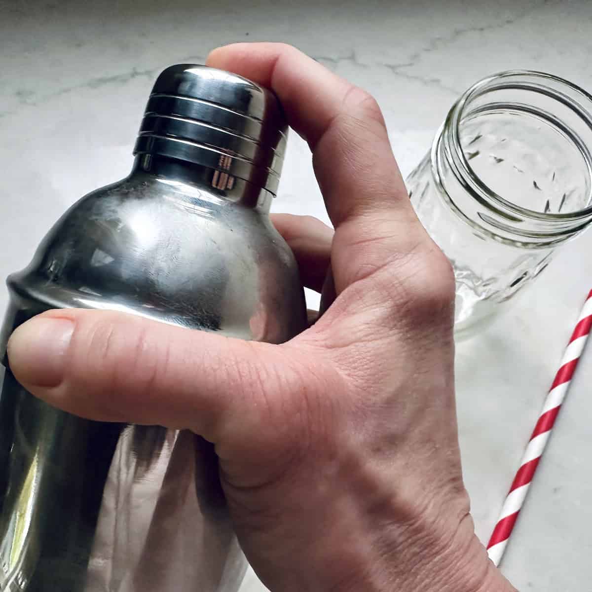 A hand holding a metal cocktail shaker next to an empty glass jar and a red-and-white striped straw on a light surface.
