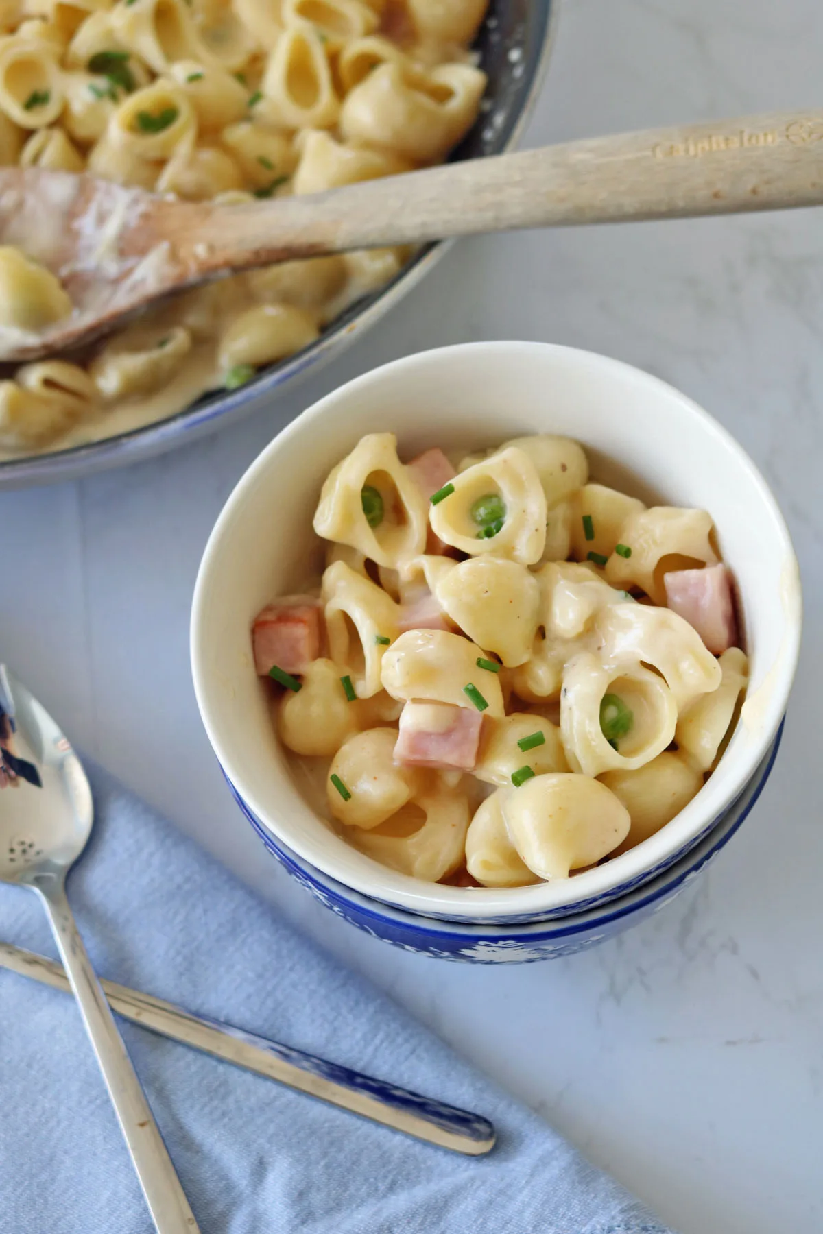 A bowl of cheesy shell pasta with diced ham and chopped chives sits on a blue napkin, next to a spoon and fork. A larger serving dish of the same pasta is in the background with a wooden spoon.