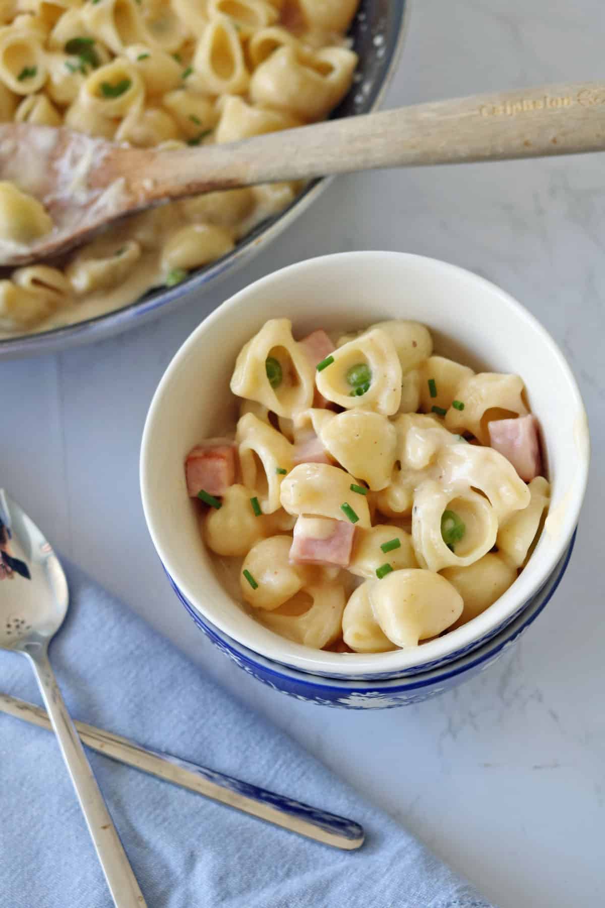 A bowl of cheesy shell pasta with diced ham and chopped chives sits on a blue napkin, next to a spoon and fork. A larger serving dish of the same pasta is in the background with a wooden spoon.