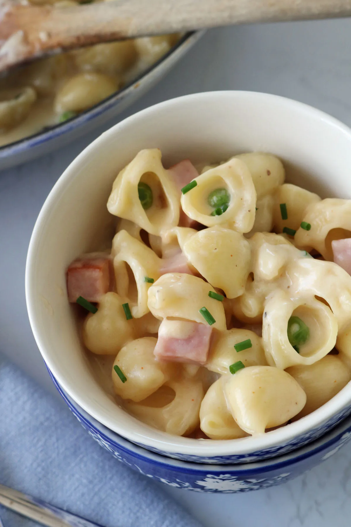 A bowl of creamy macaroni and cheese with cubes of ham and chopped chives, placed on a white surface with a blue napkin nearby and a wooden spoon in the background.
