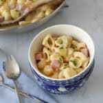 A bowl of creamy shell pasta with ham cubes and chopped chives, placed on a light blue napkin with a fork and spoon; a pan of the same pasta is in the background with a wooden spoon.