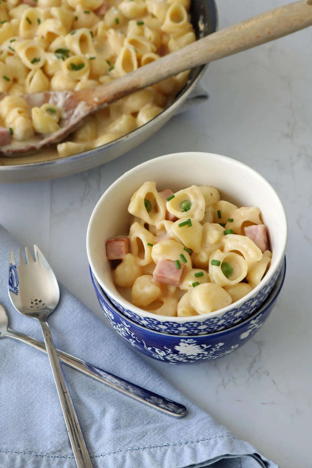 A bowl of creamy shell pasta with ham cubes and chopped chives, placed on a light blue napkin with a fork and spoon; a pan of the same pasta is in the background with a wooden spoon.