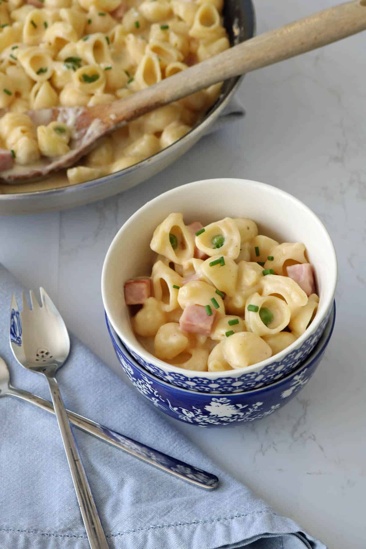 A bowl of creamy shell pasta with ham cubes and chopped chives, placed on a light blue napkin with a fork and spoon; a pan of the same pasta is in the background with a wooden spoon.