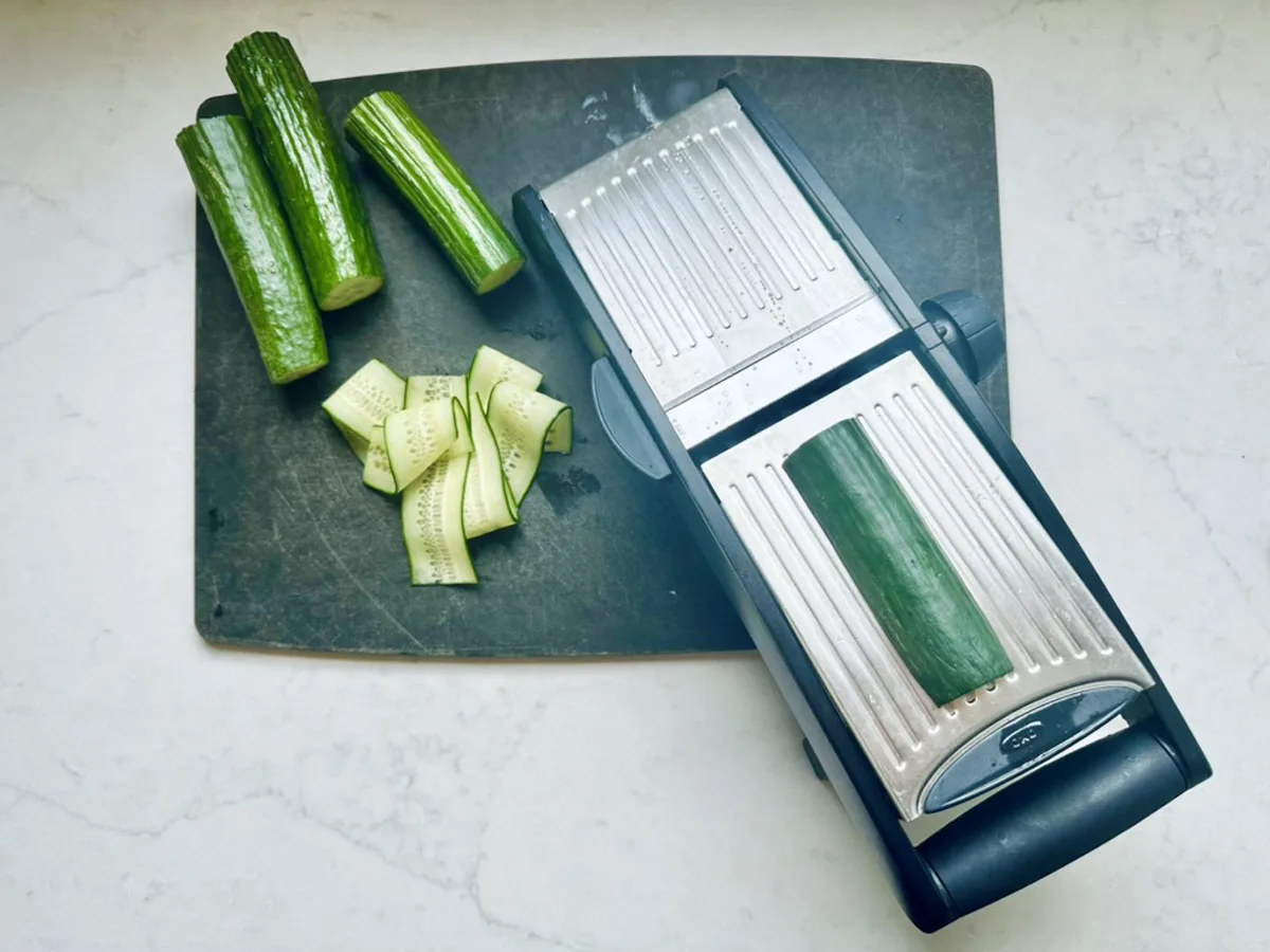 Cucumber pieces and slices are on a dark cutting board next to a mandoline slicer with a cucumber on it, ready for making Asian Cucumber Ribbon Salad, all set on a light countertop.