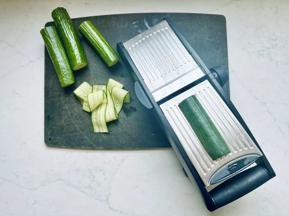 Cucumber pieces and slices are on a dark cutting board next to a mandoline slicer with a cucumber on it, ready for making Asian Cucumber Ribbon Salad, all set on a light countertop.