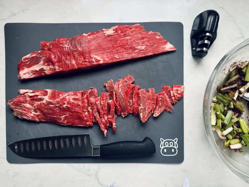 Raw flank steak is shown on a black cutting board, partially sliced into strips. A chef’s knife and meat tenderizer are nearby, with a bowl of chopped vegetables at the side.