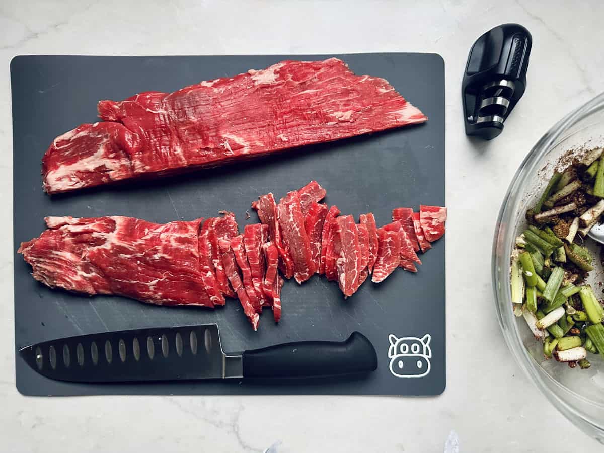 Raw flank steak is shown on a black cutting board, partially sliced into strips. A chef’s knife and meat tenderizer are nearby, with a bowl of chopped vegetables at the side.