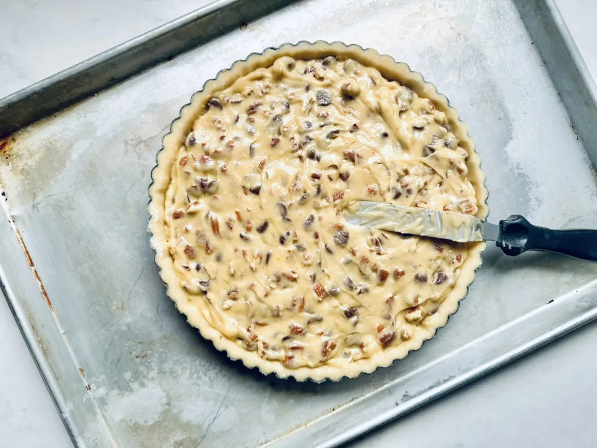 A round pie pan filled with unbaked batter containing chopped nuts sits on a metal baking sheet. A spatula covered in batter rests on the edge of the pan.