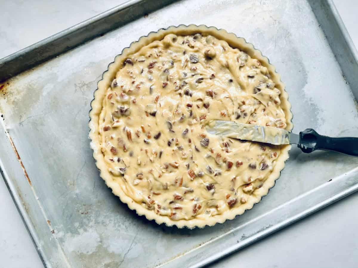 A round pie pan filled with unbaked batter containing chopped nuts sits on a metal baking sheet. A spatula covered in batter rests on the edge of the pan.
