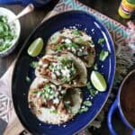 Three tacos filled with shredded meat, chopped onions, cilantro, and cheese are served on a blue oval plate with lime wedges. Surrounding dishes include rice, beans, fresh cilantro, and a bottle of green hot sauce.