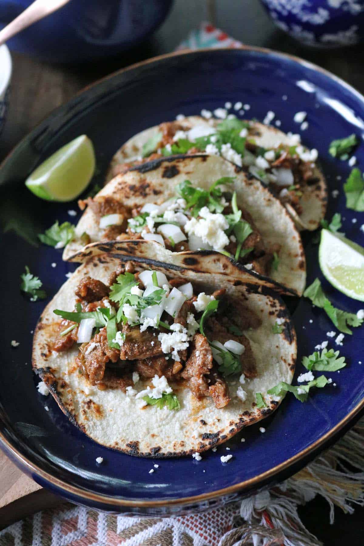 Three grilled tacos filled with seasoned meat, chopped onions, fresh cilantro, and crumbled cheese are served on a dark blue plate with lime wedges on the side.