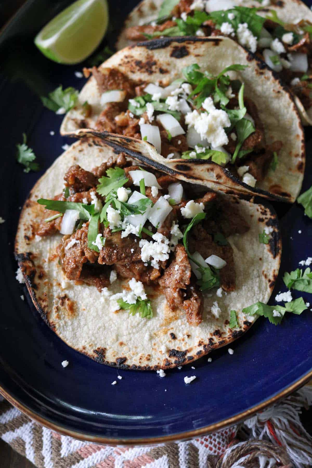 Three tacos filled with seasoned meat, topped with chopped onions, fresh cilantro, and crumbled cheese sit on a blue plate, with a wedge of lime in the background.