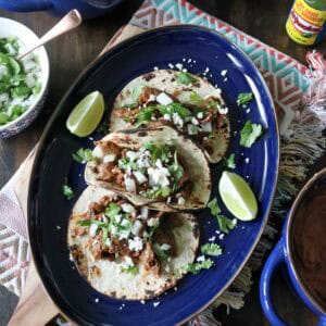Three tacos filled with shredded meat, chopped onions, cilantro, and cheese are served on a blue oval plate with lime wedges. Surrounding dishes include rice, beans, fresh cilantro, and a bottle of green hot sauce.
