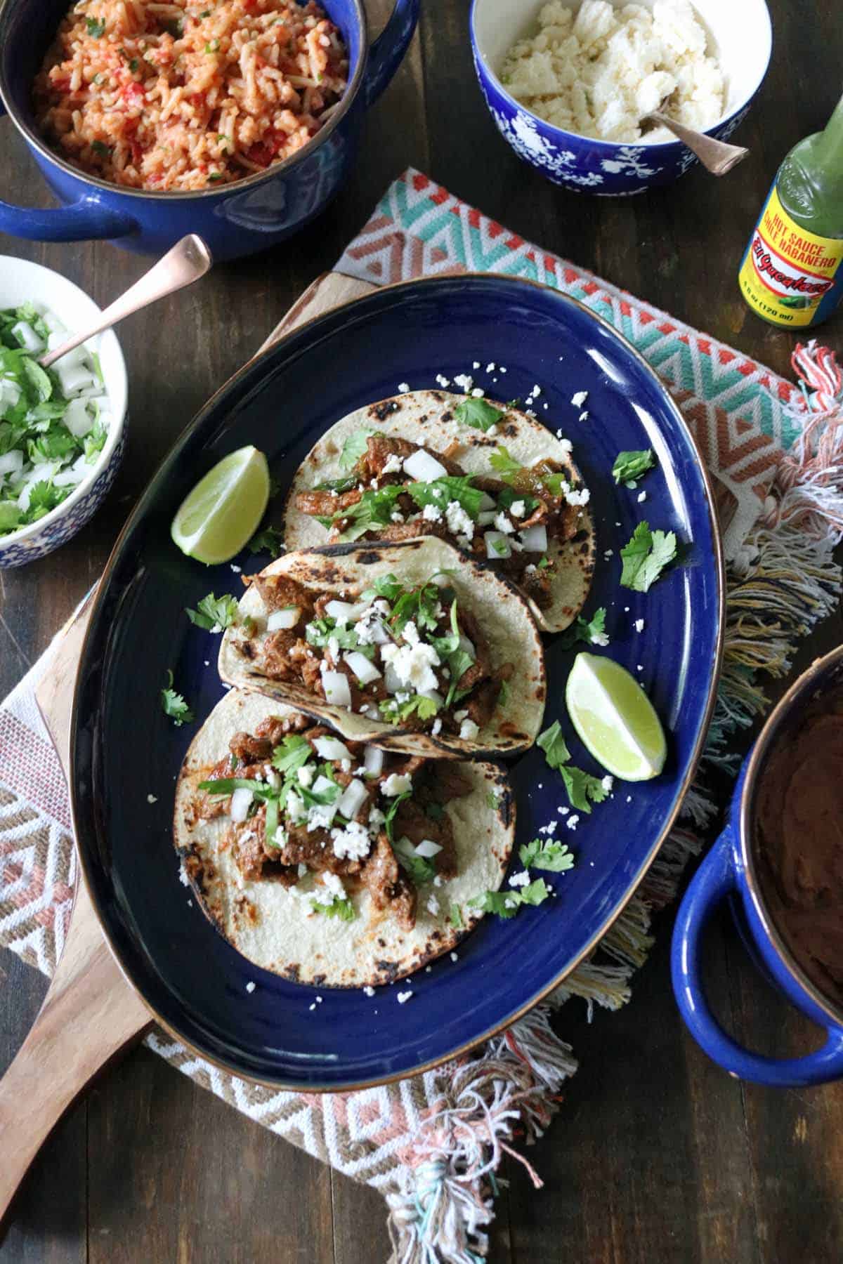 Three tacos filled with shredded meat, chopped onions, cilantro, and cheese are served on a blue oval plate with lime wedges. Surrounding dishes include rice, beans, fresh cilantro, and a bottle of green hot sauce.