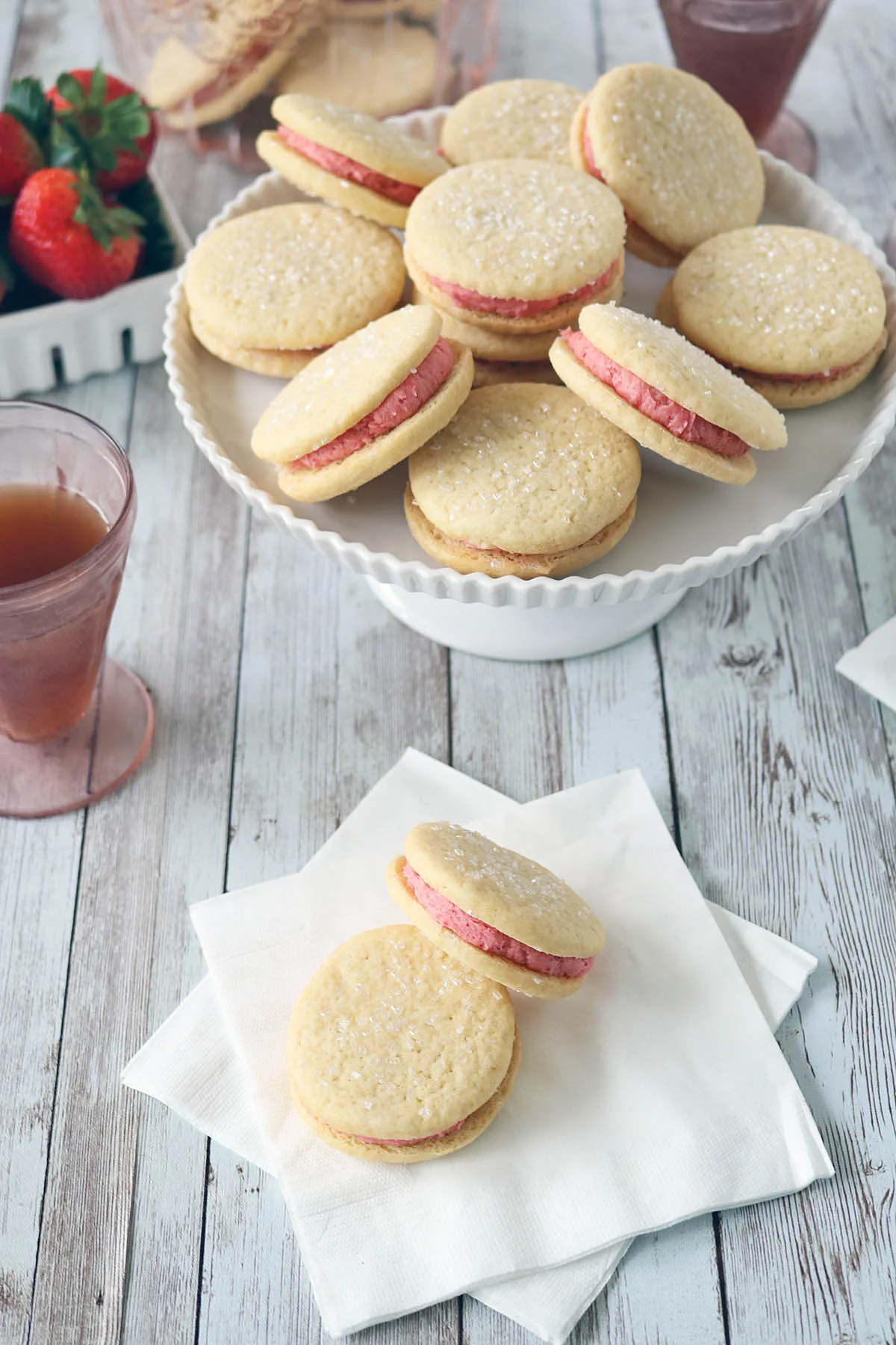 A plate of Strawberry Shortcake Cookies with pink filling and sugar sprinkles sits on a white wooden table. Two cookies are placed on napkins nearby, with drinks and fresh strawberries in the background.