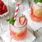 A mason jar filled with iced strawberry lemonade, reminiscent of a Strawberry Mint Julep, garnished with a lemon slice, strawberry, and mint sprig, with a red-and-white striped straw. Another drink and a bowl of strawberries are in the background.