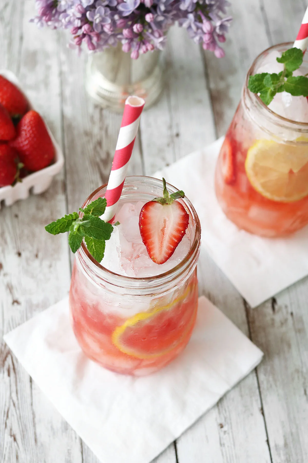 A mason jar filled with iced strawberry lemonade, reminiscent of a Strawberry Mint Julep, garnished with a lemon slice, strawberry, and mint sprig, with a red-and-white striped straw. Another drink and a bowl of strawberries are in the background.