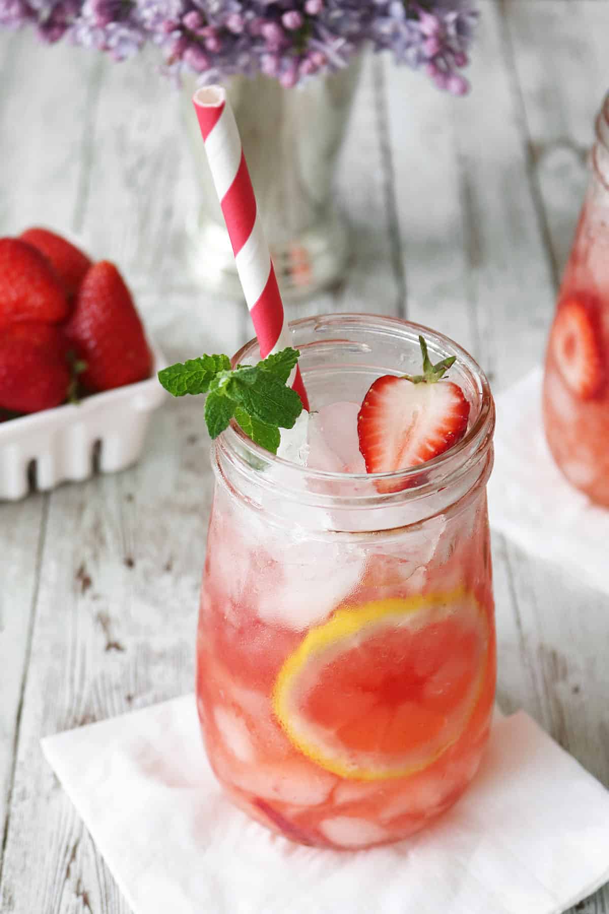 A mason jar filled with iced Strawberry Mint Julep lemonade, garnished with a lemon slice, strawberry, and mint sprig, with a red and white striped straw. A container of strawberries and flowers are in the background.