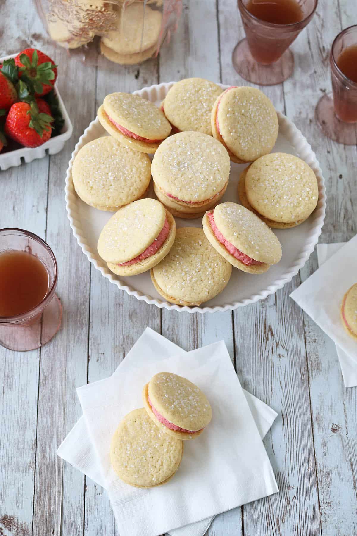 A plate of Strawberry Shortcake Cookies with pink filling sits on a wooden table, surrounded by glasses of pink drink, fresh strawberries, and napkins with additional cookies.