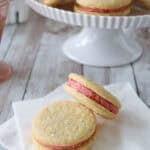 Two Strawberry Shortcake Cookies with pink filling rest on white napkins, while more of these sparkling sugar-topped cookies are displayed on a white cake stand in the background.